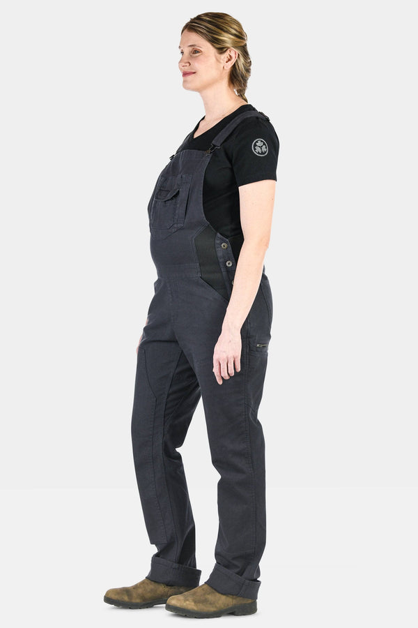 Side view of a woman wearing Dovetail Workwear's Freshley Maternity Overalls in Navy Stretch Canvas with reinforced knees and flex suspenders