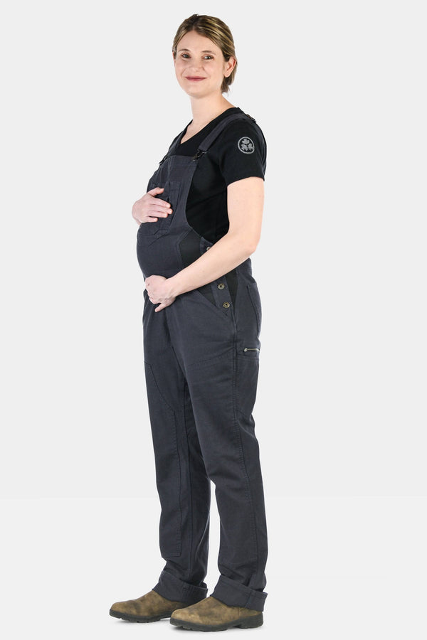 Pregnant woman wearing Dovetail Workwear Freshley Maternity Overalls in Navy Stretch Canvas with black shirt and brown boots, hands on belly, side view