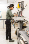 Woman wearing Dovetail Workwear Maven X Graphene Denim Pant in Black, demonstrating the slim-fit carpenter pants while operating machinery in a workshop setting