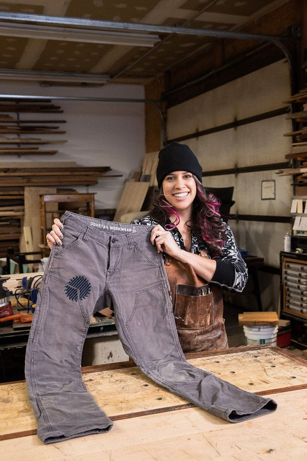 Woman holding up grey Dovetail Workwear pants with visible Black Denim Pocket Patch in a workshop setting, demonstrating DIY repair patch application