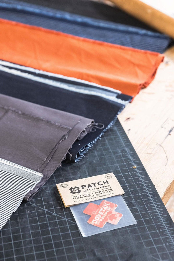 Dovetail Workwear x NoSo DIY Repair Patches displayed on a cutting mat, showing multiple fabric patches in paprika orange, indigo denim, black, and grey canvas with adhesive backing