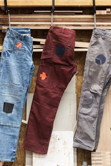 Three pairs of work pants hanging on a wooden rack display Dovetail Workwear x NoSo DIY Repair Patches in Paprika Orange and Grey Canvas, applied to denim and canvas in various sizes