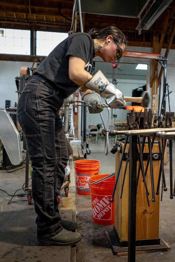 Woman wearing Dovetail Workwear Old School High Rise Graphene Denim Pant in Heather Black, working at anvil in shop