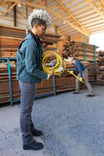 Woman wearing Dovetail Workwear Old School Trucker Canvas Work Jacket in Lodge Green, holding a yellow extension cord in an industrial warehouse setting