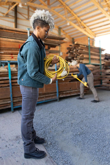 Woman wearing Dovetail Workwear Old School Trucker Canvas Work Jacket in Lodge Green, holding a yellow extension cord in an industrial warehouse setting