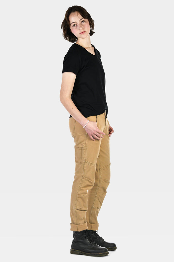 Side view of a woman wearing Dovetail Workwear GO TO™ Stretch Canvas Pant in Sawdust Brown with visible nested pockets and cuffed inseam, paired with a black v-neck tee and black boots