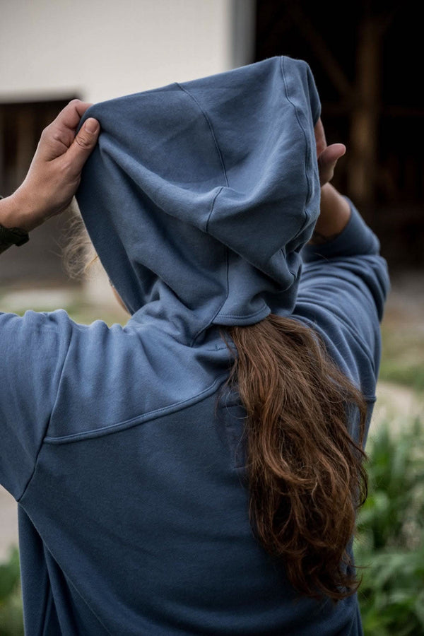 Back view of a woman wearing the John Deere X Dovetail Workwear Early Bird Hoodie in Steel Blue, showing the ponytail slit and cowl neck design