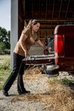 Woman wearing John Deere X Dovetail Workwear Into Town Bootcut Jeans in Classic Black while working near a truck bed, demonstrating the mid-rise fit and bootcut silhouette