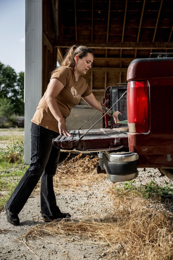 Woman wearing John Deere X Dovetail Workwear Into Town Bootcut Jeans in Classic Black while working near a truck bed, demonstrating the mid-rise fit and bootcut silhouette