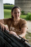 Woman wearing the John Deere X Dovetail Workwear Heartland T-Shirt in Sawdust Brown, leaning against a fence with farmland visible in the background