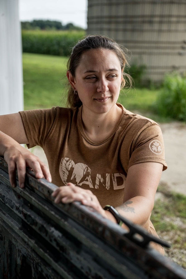 Woman wearing the John Deere X Dovetail Workwear Heartland T-Shirt in Sawdust Brown, leaning against a fence with farmland visible in the background