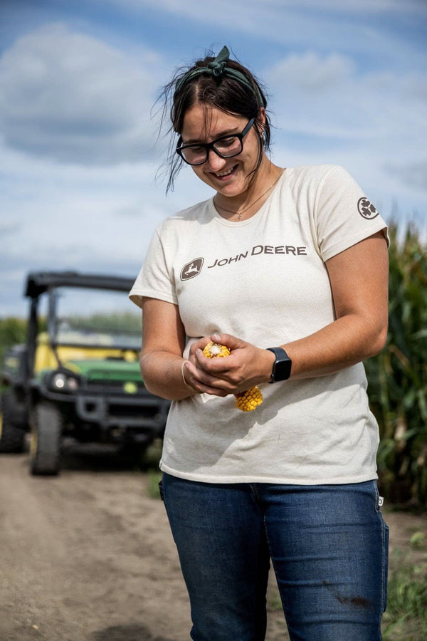 Woman wearing the John Deere X Dovetail Workwear T-Shirt in Heathered Ecru White, holding corn in a farm field with equipment visible behind her