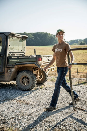 Woman wearing John Deere X Dovetail Workwear Into Town Bootcut Jeans in Indigo Denim, standing by a farm gate with a John Deere vehicle in the background