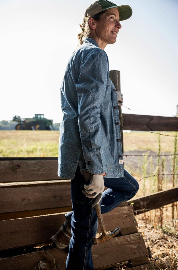 Back view of a woman wearing the John Deere X Dovetail Cultivator Work Shirt in Indigo Blue Dobby, standing at a ranch fence overlooking farmland