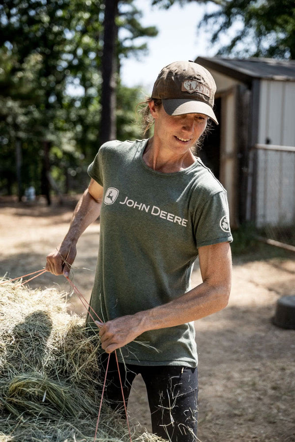 Woman wearing the Heathered Green John Deere X Dovetail Workwear T-Shirt while handling hay, smiling at the camera in a farm setting