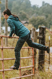 Woman climbing a rusty gate in Dovetail Workwear's Britt X Pant Power Hemp in Indigo, demonstrating the durable hemp fabric's flexibility and reinforced construction for active work and outdoor tasks