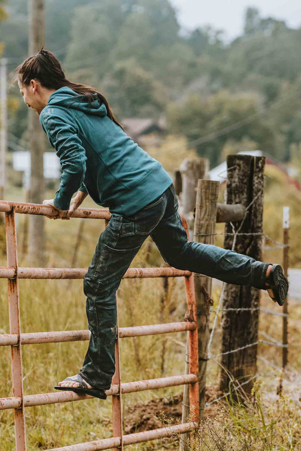 Woman climbing a rusty gate in Dovetail Workwear's Britt X Pant Power Hemp in Indigo, demonstrating the durable hemp fabric's flexibility and reinforced construction for active work and outdoor tasks