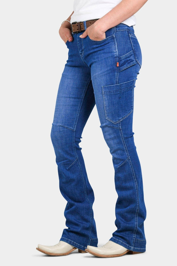 Woman wearing Dovetail Workwear DX Bootcut Pant in Blue Sky Denim, featuring visible tool loop, nested pockets, and articulated knee design, paired with white boots