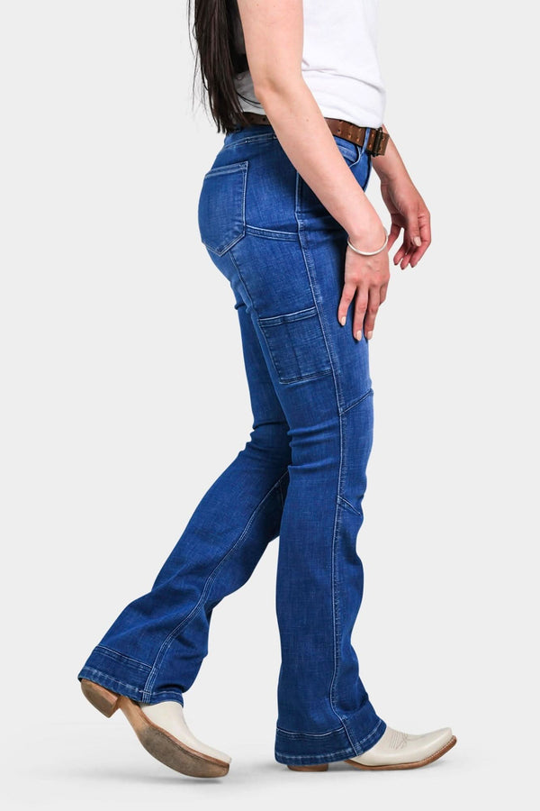 Side view of a woman wearing Dovetail Workwear's DX Bootcut Pant in Blue Sky Denim, showcasing the bootcut silhouette, nested pockets, and tool loop detail