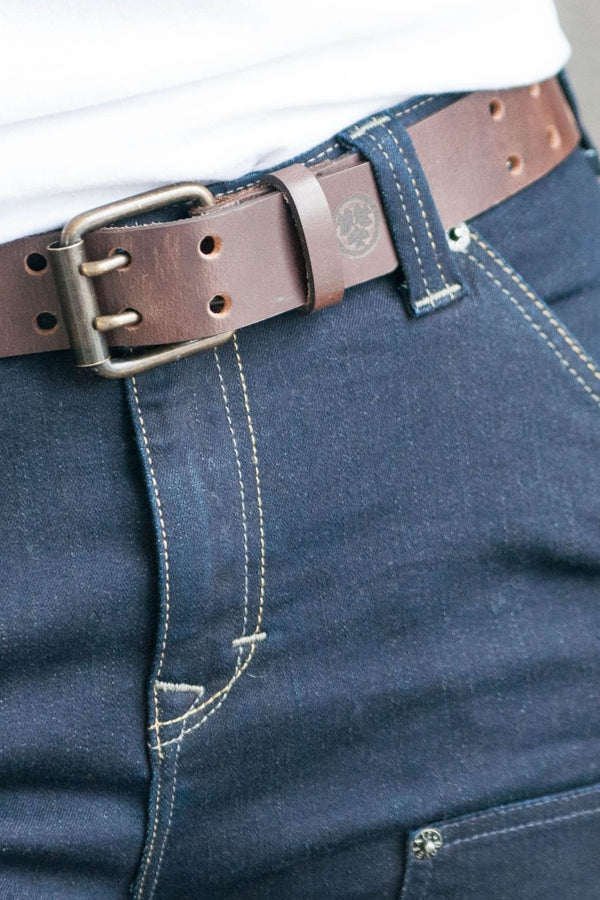 Detail view of the Double Pronged Work Belt by Dovetail Workwear in Brown Clay leather, showing the double-pronged buckle and full-grain buffalo leather construction on dark denim jeans