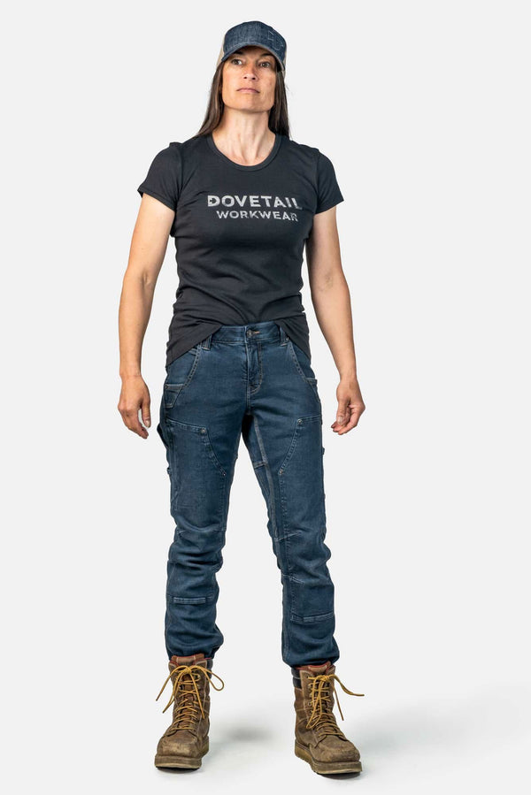 Woman wearing Dovetail Workwear's Britt X Pant Power Hemp in Indigo with reinforced knees, paired with a black logo tee and denim cap
