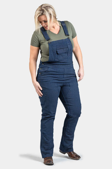 Freshley Overalls in Wabash Stripe by Dovetail Workwear worn by a woman with blonde hair, featuring pinstriped bib overalls with adjustable suspenders and reinforced knees, paired with an olive green v-neck t-shirt and brown shoes