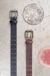 Double Pronged Work Belt by Dovetail Workwear displayed in Black Tar and Brown Clay with double-pronged buckles and circular leather swatches on textured background