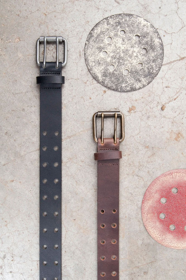 Double Pronged Work Belt by Dovetail Workwear displayed in Black Tar and Brown Clay with double-pronged buckles and circular leather swatches on textured background