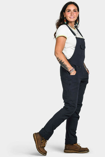 Side view of a woman wearing Dovetail Workwear's Freshley Drop Seat Overalls in Navy Canvas with brown work boots and a white t-shirt