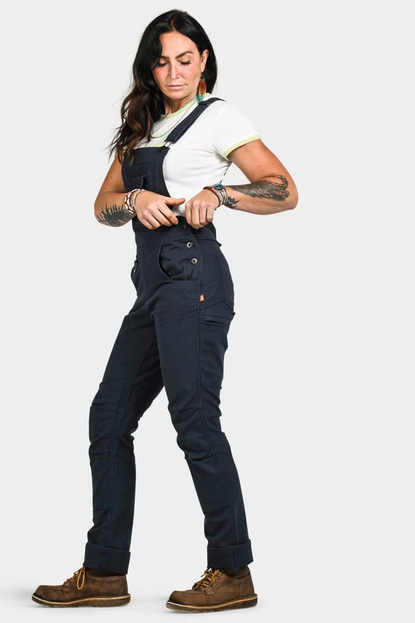 Woman wearing Dovetail Workwear Freshley Drop Seat Overalls in Navy Canvas with white t-shirt and brown work boots, showcasing the reinforced knees and tool loops