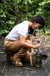 Woman crouching in a garden wearing Dovetail Workwear's Maven X Pant in Saddle Brown Canvas, demonstrating the reinforced knees and tool loop while working outdoors