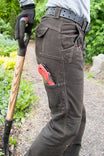 Back view of a woman wearing Dovetail Workwear's Day Construct Pant in Brown Canvas, featuring reinforced knees, phone pocket, and a leather belt, while holding a work tool