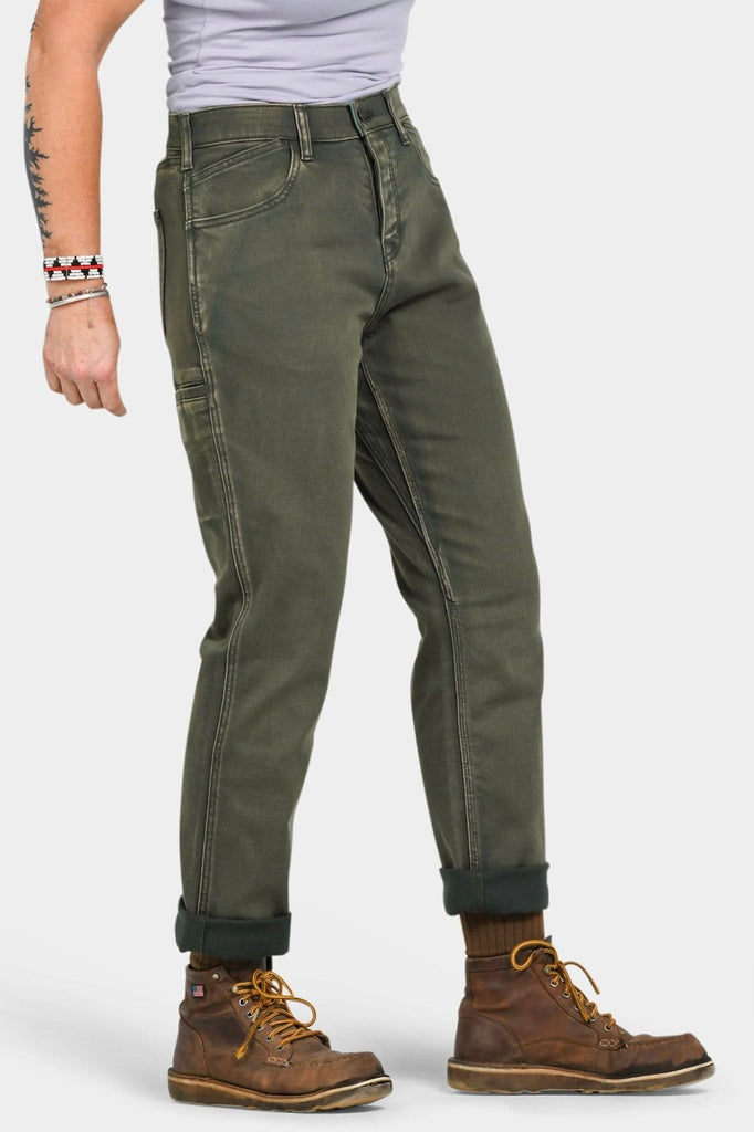 Side view of a woman wearing Dovetail Workwear's Shop Pant in Olive Green Denim, cuffed at the ankles, paired with brown work boots