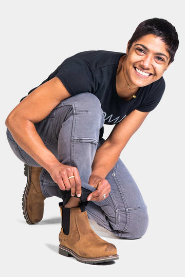 Woman kneeling while adjusting the cuff of grey Britt Utility Pants in Stretch Thermal Denim by Dovetail Workwear, showcasing the reinforced knees and straight leg fit