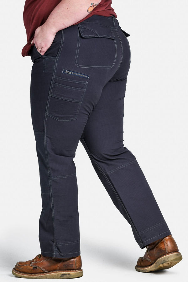 Side view of a woman wearing Dovetail Workwear's Day Construct Pant in Lightweight Dark Navy Ripstop, showcasing the phone pocket, reinforced knees, and relaxed fit with brown boots