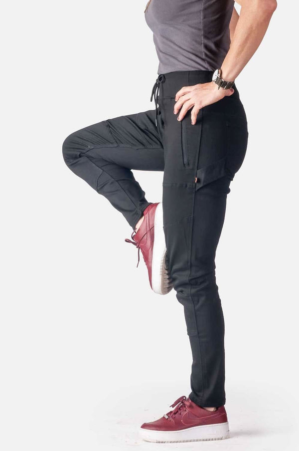 Woman wearing Dovetail Workwear Christa DIY Pant in Black Performance Modal, shown lifting one leg to display the reinforced knee and slim leg fit in a dynamic pose