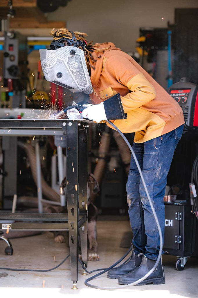 Woman welding in Dovetail Workwear's Christa DIY Pant in Sulfur Navy, showcasing reinforced knees and tool loop functionality in an industrial workshop setting