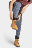 Woman wearing Dovetail Workwear Maven X Slim Pant in Grey Stretch Thermal Denim with tan lace-up boots, adjusting black sock cuff, showcasing reinforced knee and slim fit