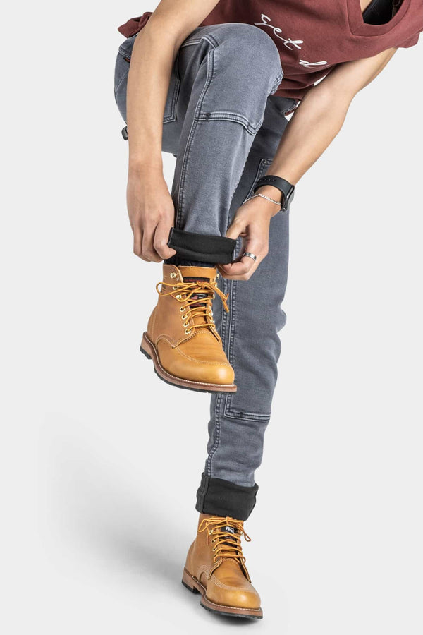 Woman wearing Dovetail Workwear Maven X Slim Pant in Grey Stretch Thermal Denim with tan lace-up boots, adjusting black sock cuff, showcasing reinforced knee and slim fit