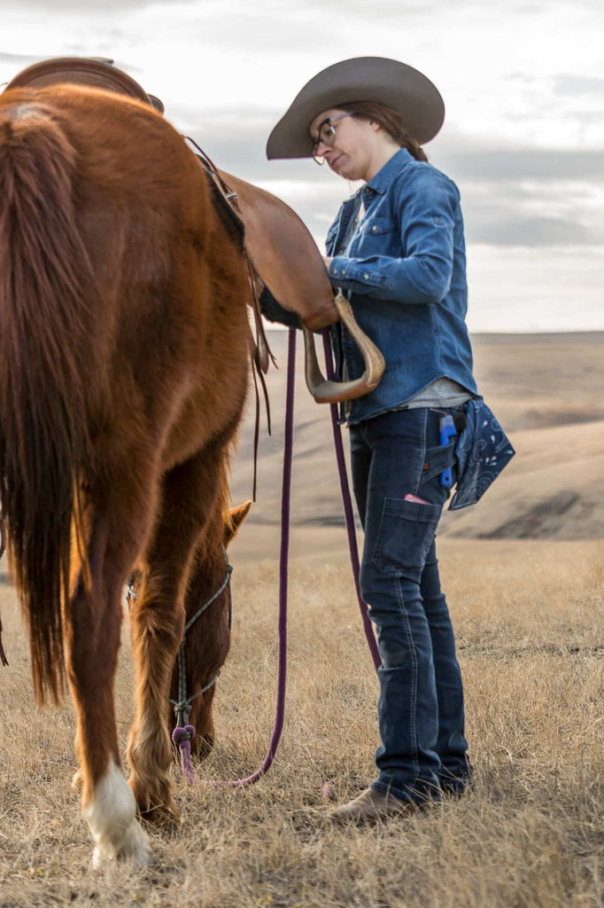 Woman wearing Dovetail Workwear's DX Bootcut Pant in CORDURA® Indigo Denim while saddling a brown horse on an open ranch landscape