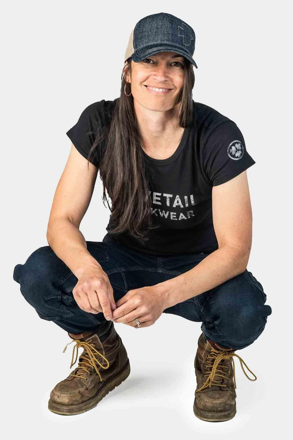 Woman wearing the Dovetail Embroidered Shop Cap in blue denim and black, crouching and smiling at the camera, paired with dark jeans and work boots