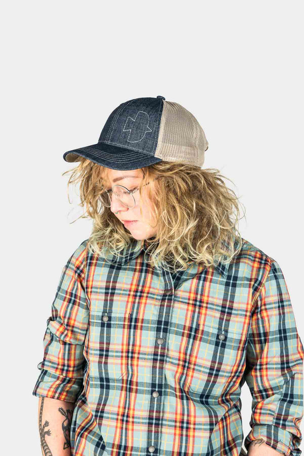Woman wearing the Dovetail Embroidered Shop Cap in Blue/Black denim with contrasting cream mesh panels, paired with a colorful plaid shirt