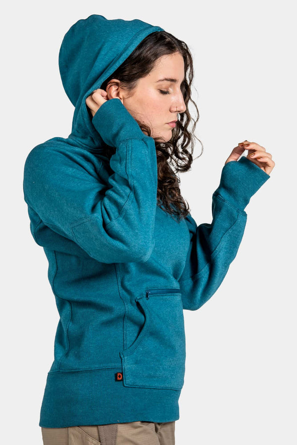 Side view of a woman wearing the Anna Pullover Hoodie in Dovetail Blue by Dovetail Workwear, featuring a hood, thumbhole cuffs, and front pockets