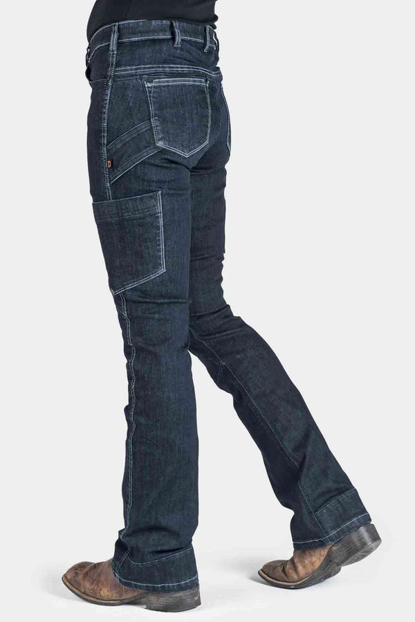 Side view of a woman wearing Dovetail Workwear's DX Bootcut Pant in CORDURA® Indigo Denim, showcasing the bootcut silhouette, phone pocket, and articulated knee design with brown boots