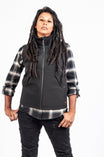 Woman wearing Dovetail Workwear's M'fon Work Vest in black over a plaid shirt, showcasing the water-resistant softshell with high collar and deep pockets