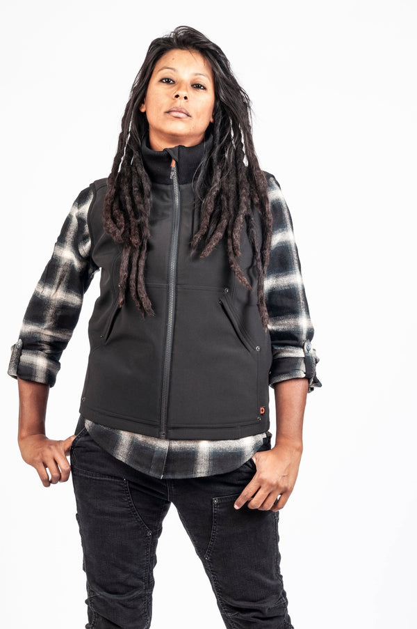Woman wearing Dovetail Workwear's M'fon Work Vest in black over a plaid shirt, showcasing the water-resistant softshell with high collar and deep pockets