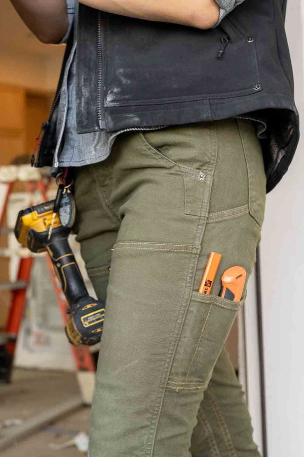 Detail view of the Maven X Pant in Moss Green Canvas by Dovetail Workwear, showing nested pockets, tool loop, and reinforced knees on a woman wearing the slim-fit work pants