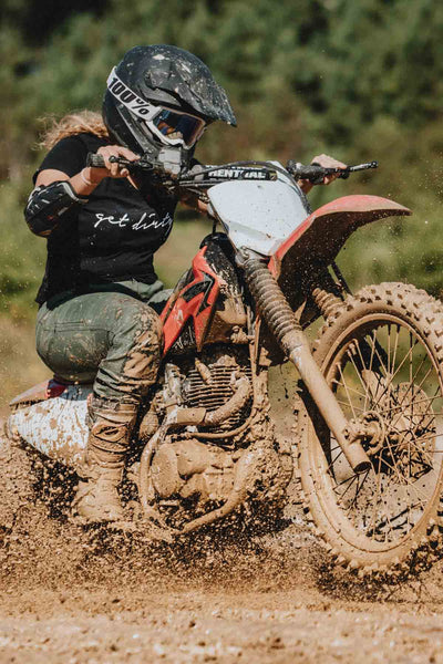 Woman in mud on dirt bike wearing Dovetail Workwear Britt X Ultralight Work Pant in Lichen Green Ripstop, demonstrating durability and water-resistant features in outdoor conditions