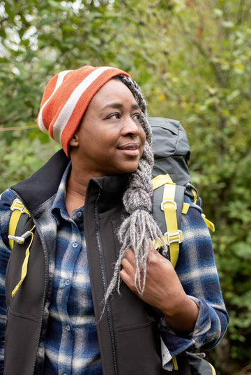 Woman wearing the Dovetail Workwear M'fon Work Vest in black, layered over a plaid shirt, standing outdoors with a backpack